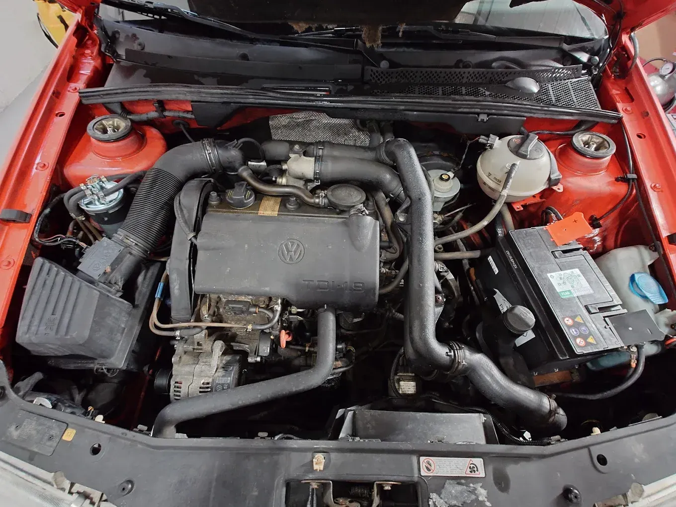 Capot de voiture rouge ouvert révélant un compartiment moteur rouge avec batterie, filtre à air et autres composants du moteur
