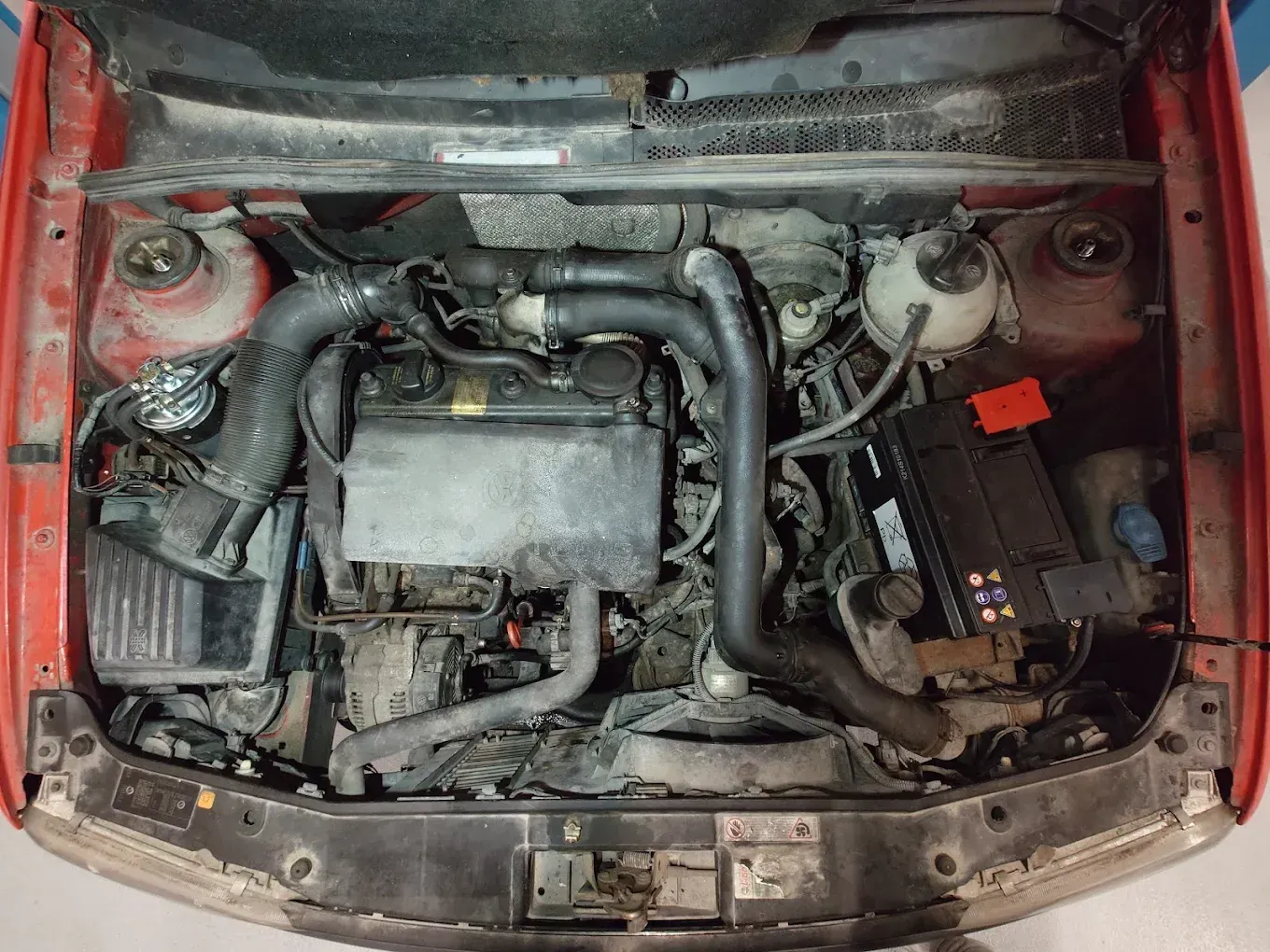 Capot d'une voiture rouge ouvert laissant apparaître le compartiment moteur, avec les durites, la batterie et le boîtier du filtre à air