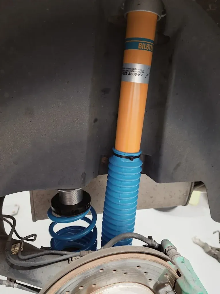 Gros plan sur la suspension arrière d'une voiture, avec un amortisseur orange et un ressort hélicoïdal bleu, sous le passage de roue