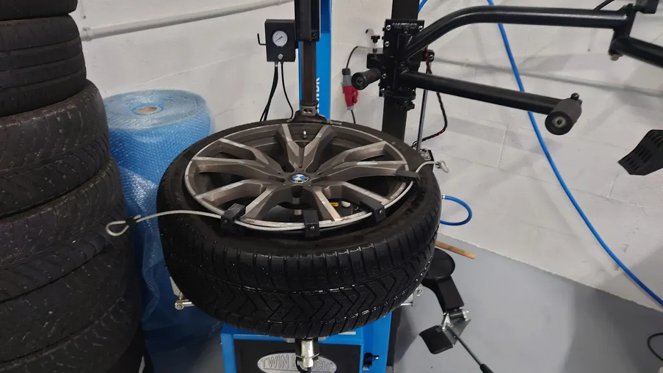 Un pneu de voiture monté sur une machine d'équilibrage dans un garage, avec des pneus empilés à proximité.