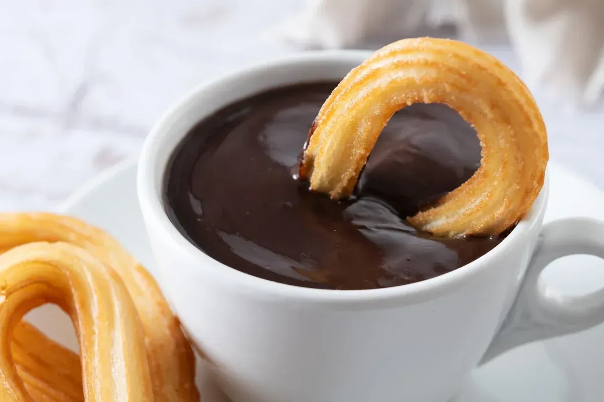Churros bañados en una taza de chocolate negro sobre una mesa blanca.