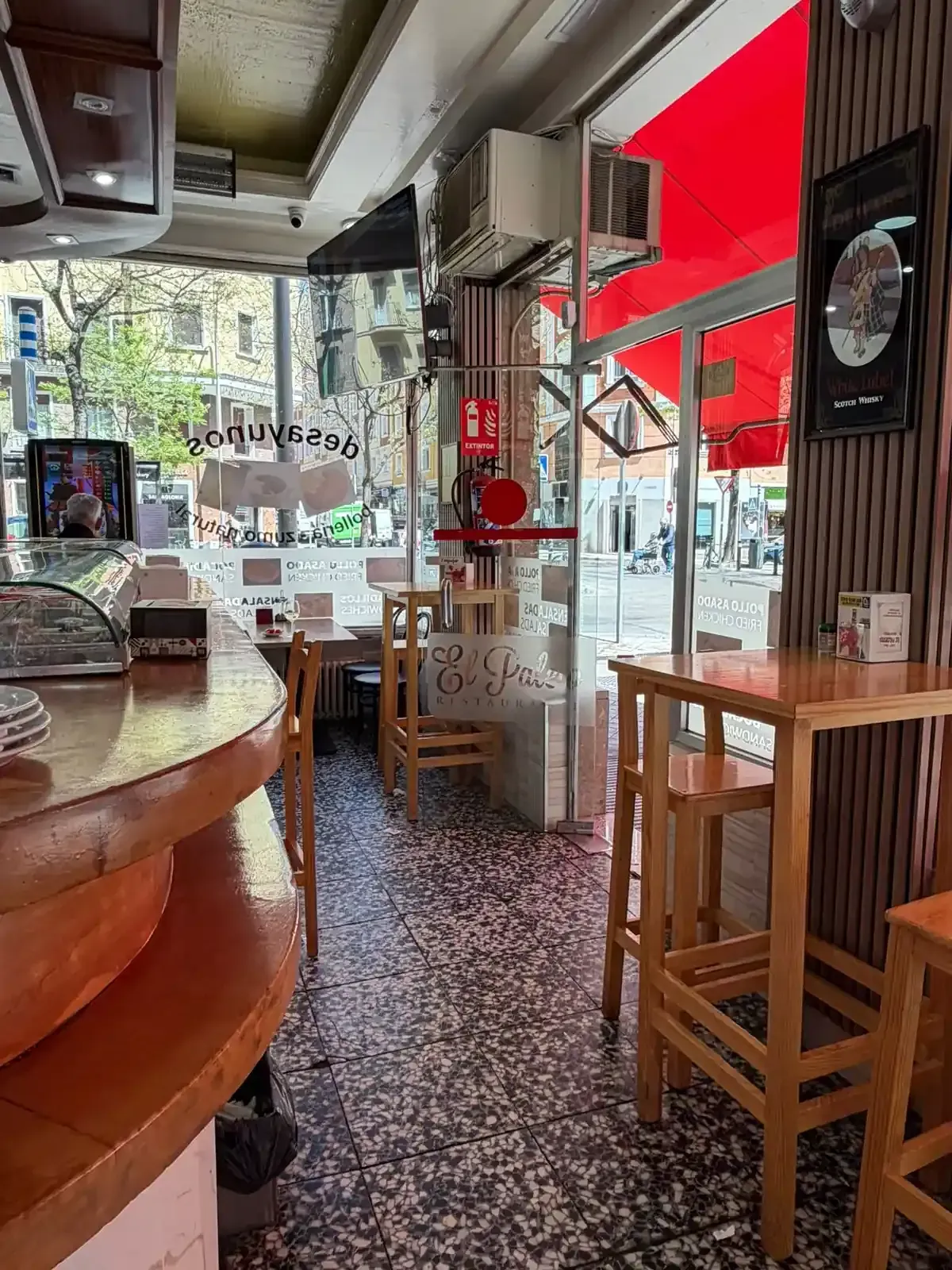 Interior acogedor de una cafetería con mesas de madera, toldo rojo y una barra junto a grandes ventanales.