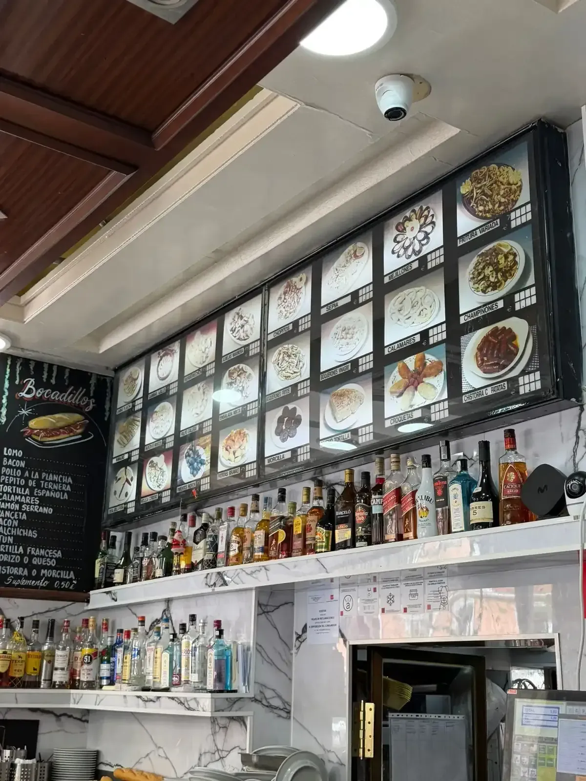 Pared con el menú del restaurante, botellas de bebidas y un mostrador bajo una claraboya.