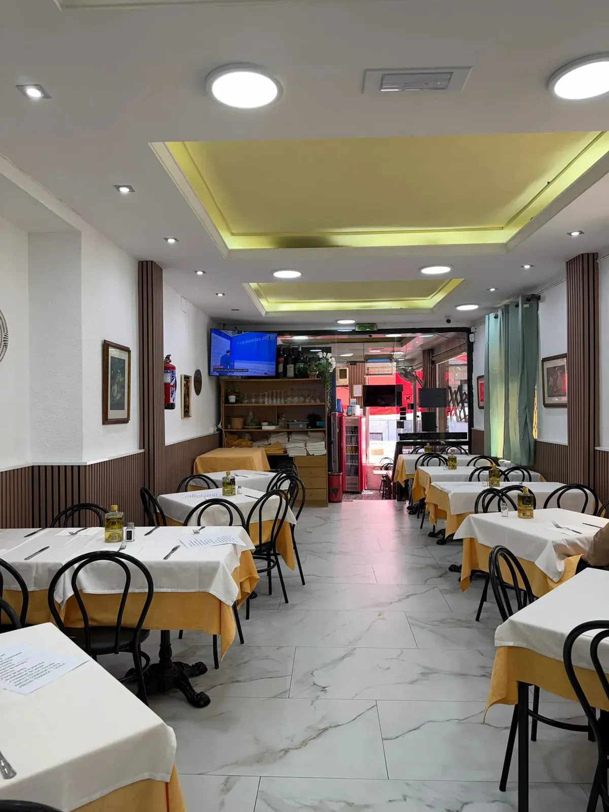Comedor vacío de un restaurante con manteles blancos, suelo de mármol y luces cálidas en el techo, visto desde la entrada.