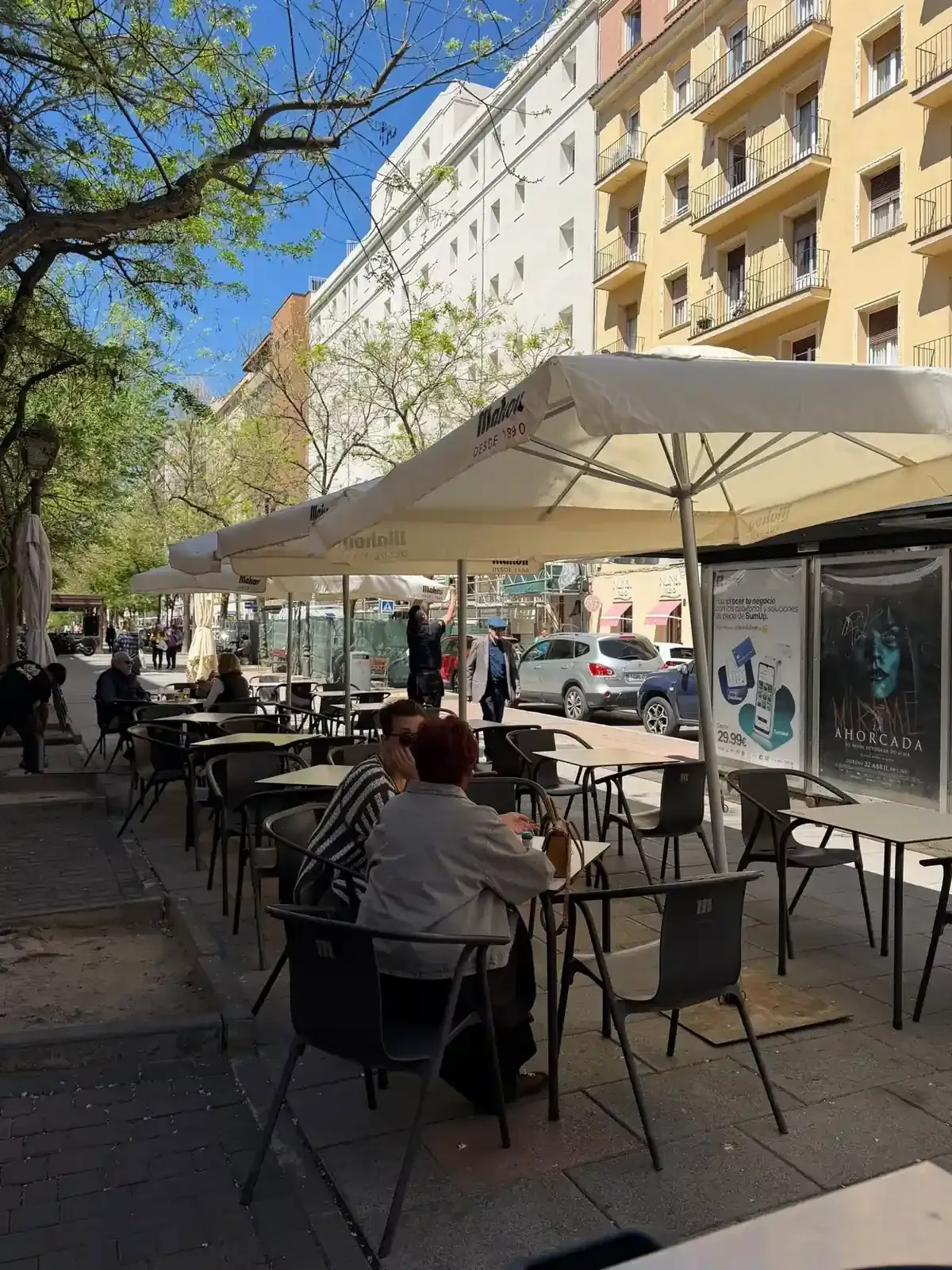 Mesas de café al aire libre bajo grandes sombrillas en una soleada calle de la ciudad, rodeada de edificios de apartamentos y árboles.