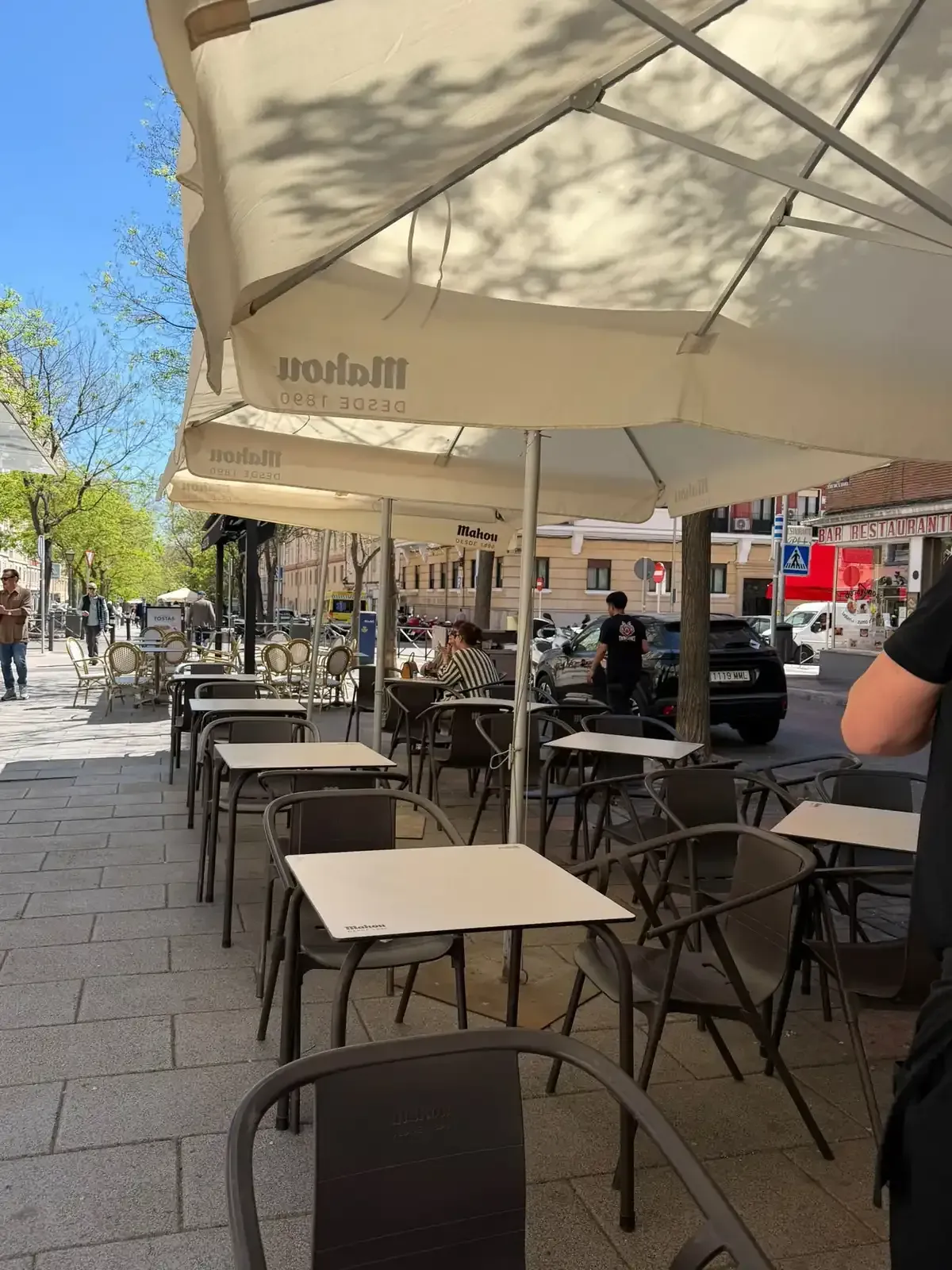 Terraza de una cafetería con mesas y sillas al aire libre, bajo sombrillas blancas, a lo largo de una acera soleada, aunque algunas están vacías.