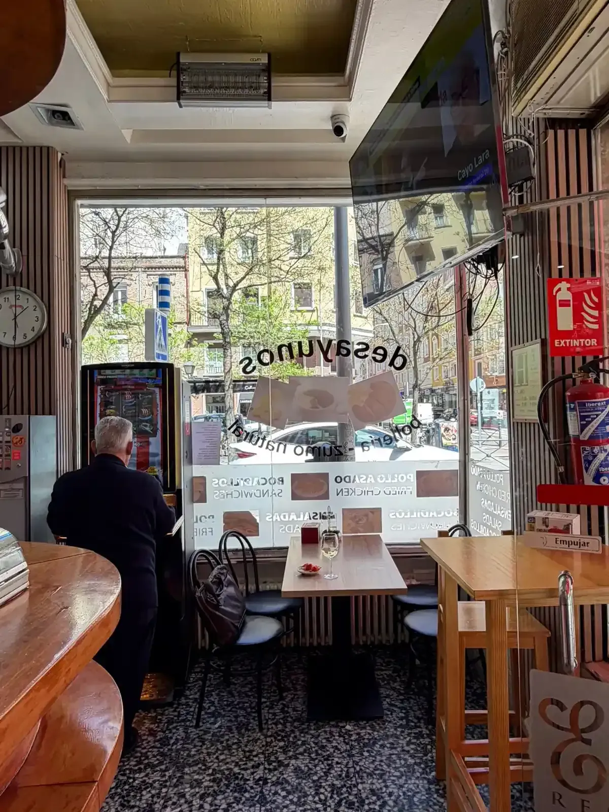 Interior acogedor de una cafetería con un cliente sentado junto a la ventana principal y vista a la calle.
