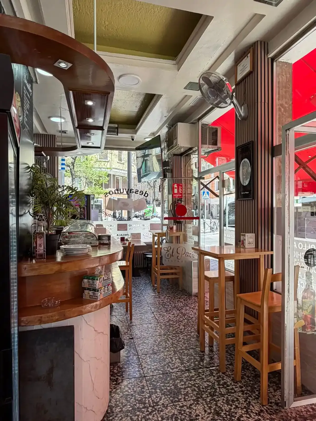 Interior estrecho de una cafetería con mesas de madera, taburetes y toldos rojos a lo largo de una luminosa ventana que da a la acera.
