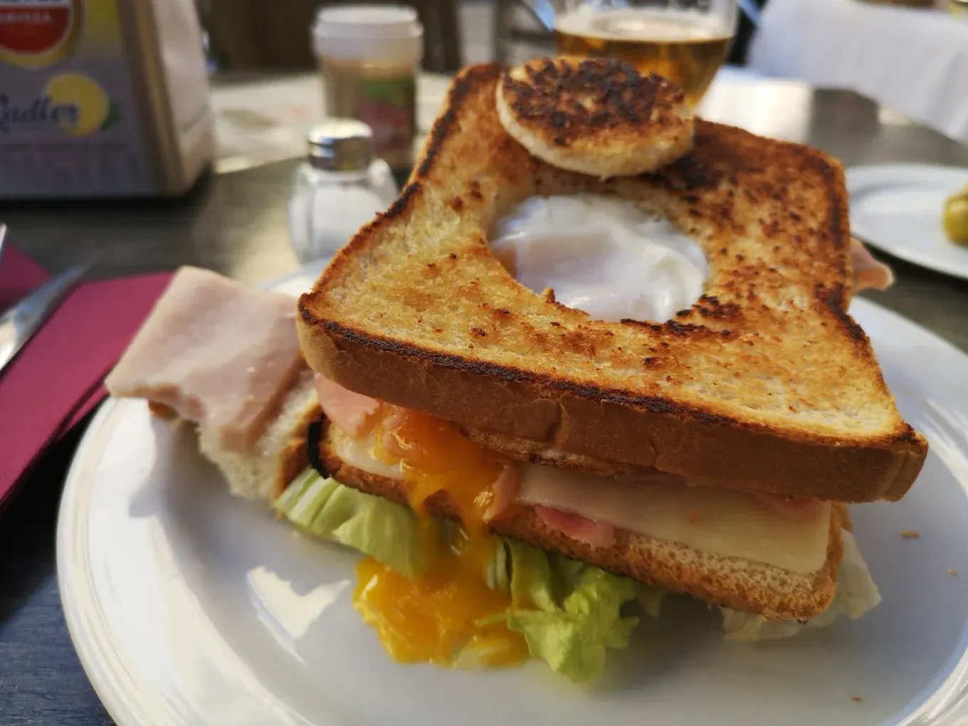 Sándwich tostado apilado en un plato blanco con lechuga, jamón y queso fundido, coronado con un huevo frito.
