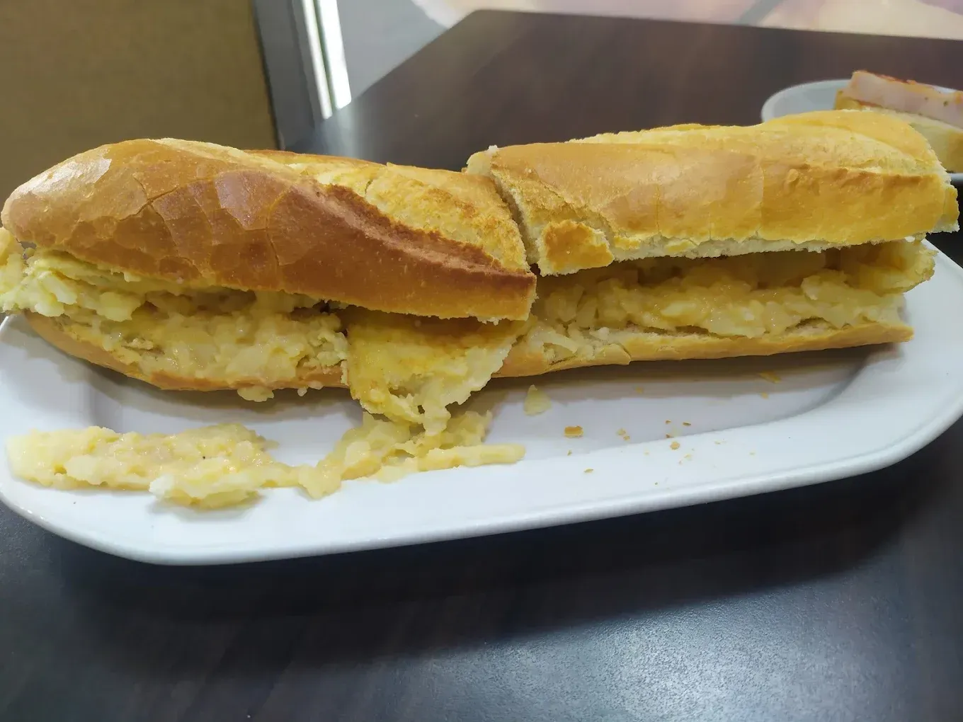 Dos mitades de un sándwich en un plato blanco, con migas sobre una mesa oscura.