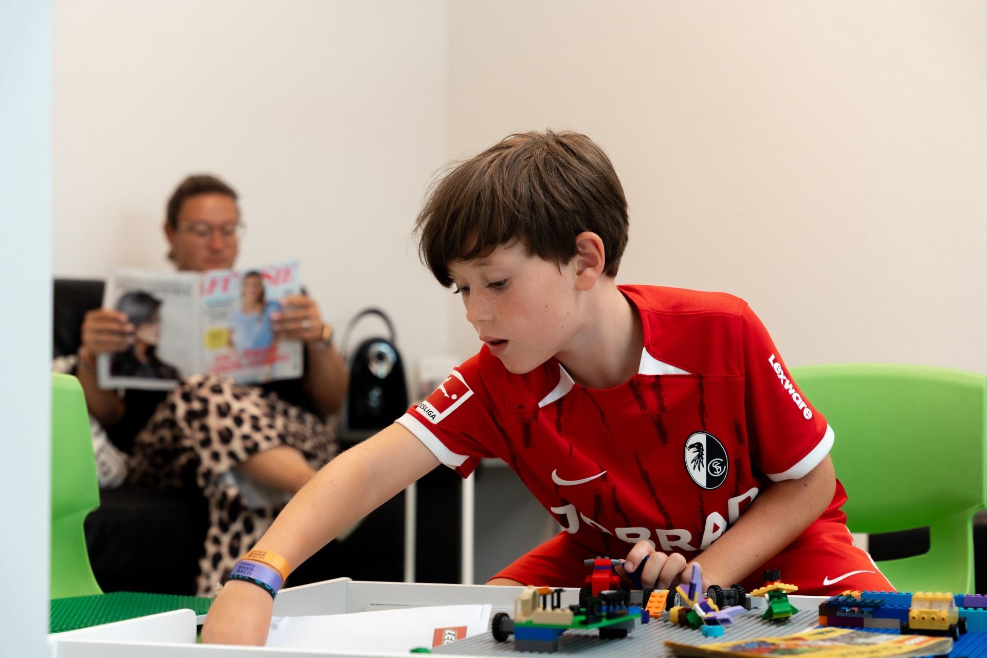 Ein Kind in einem roten Fußballtrikot spielt mit Bauklötzen an einem Tisch; im Hintergrund sitzt ein Erwachsener.