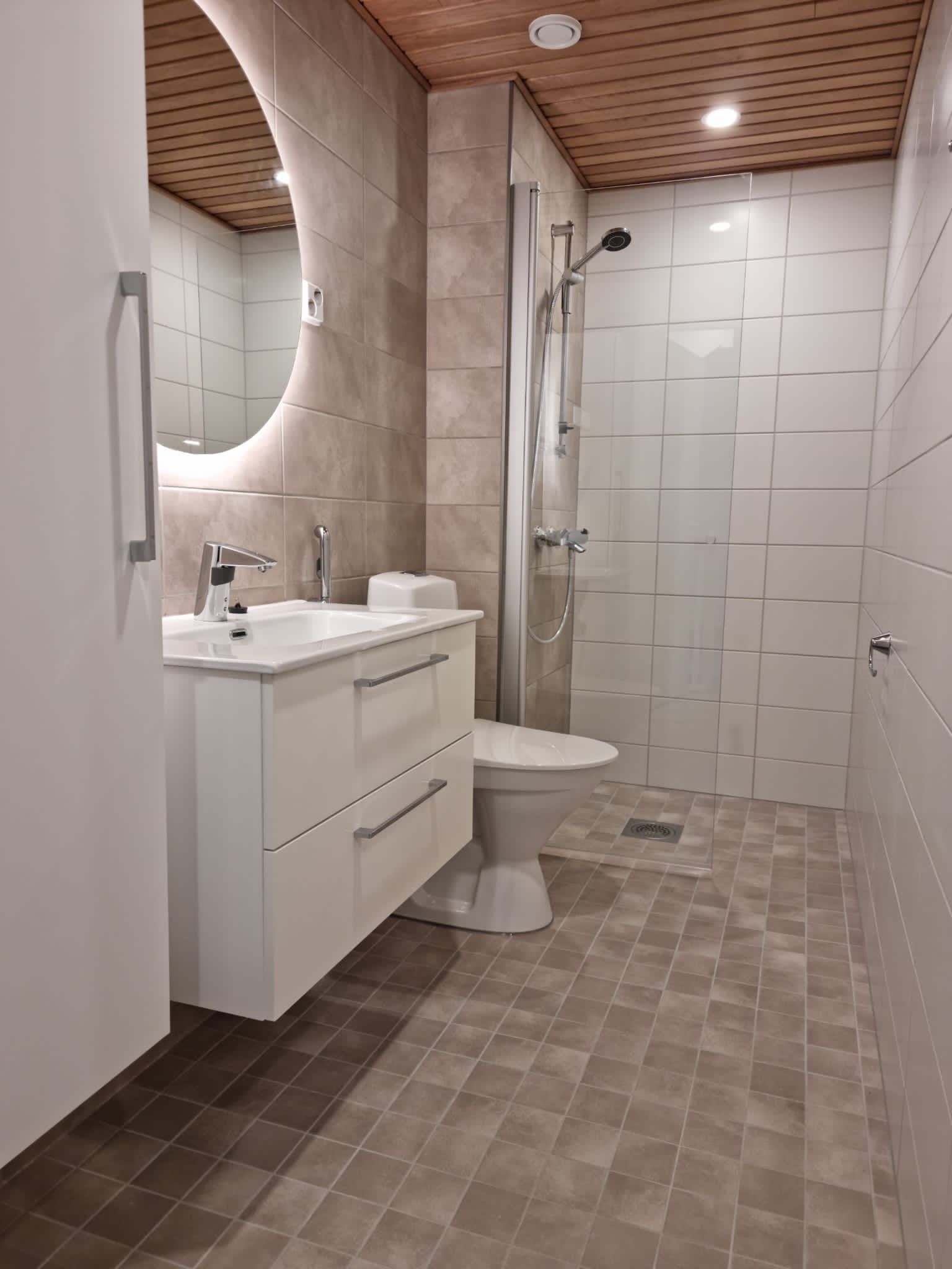 Moderni kylpyhuone, jossa valkoinen pesuallas, pyöreä peili, wc, suihku sekä laattalattia ja -seinät.
