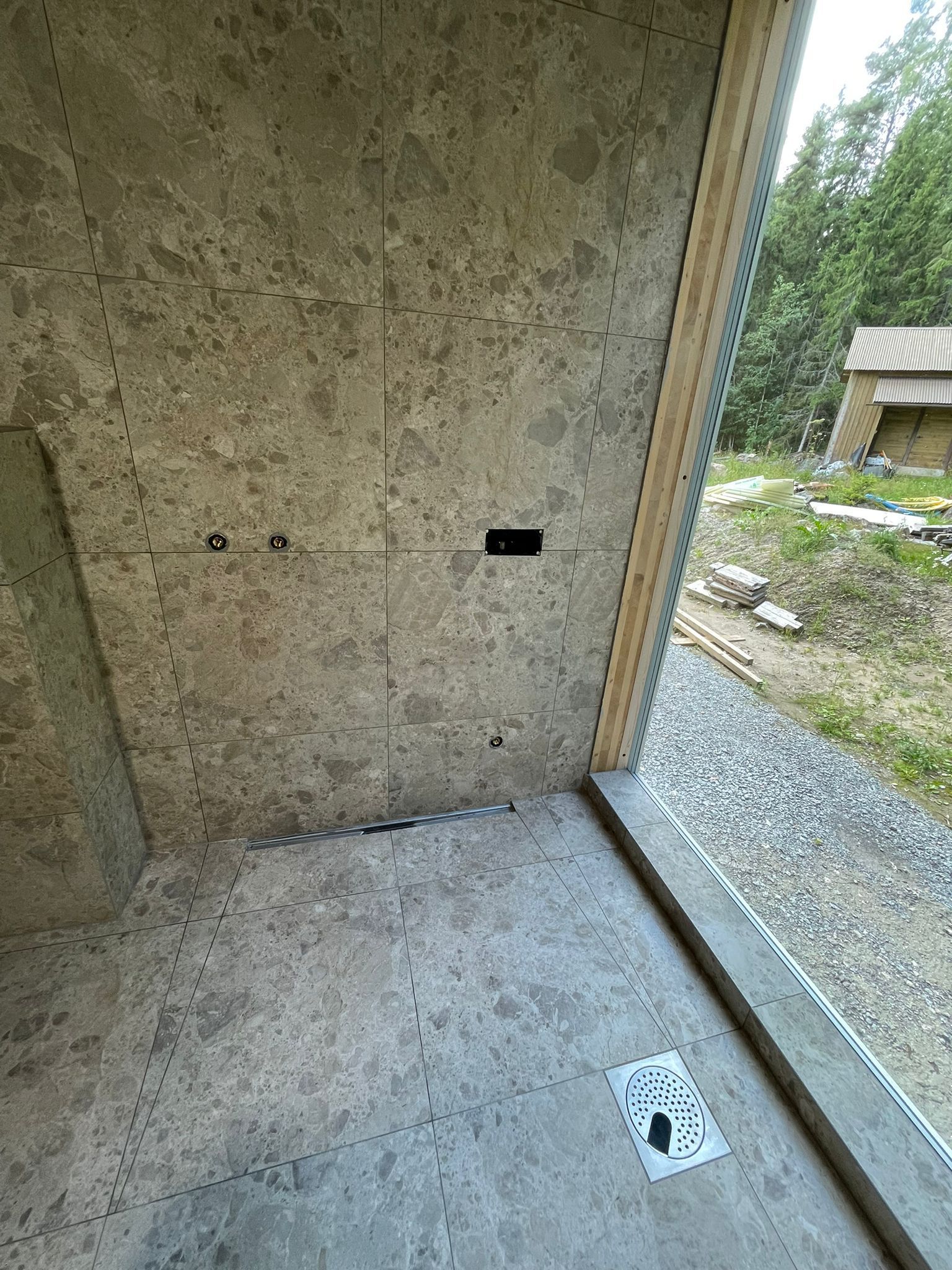 Tyhjä betonihuone, jossa on näkyvä seinä, lattiakaivo ja lasinen oviaukko sorapihalle.