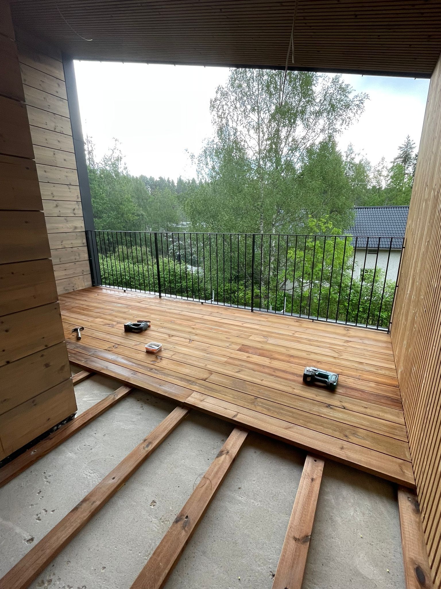 Rakenteilla oleva katettu parveke, jossa puiset lattiapalkit, musta kaide ja puita sen takana.