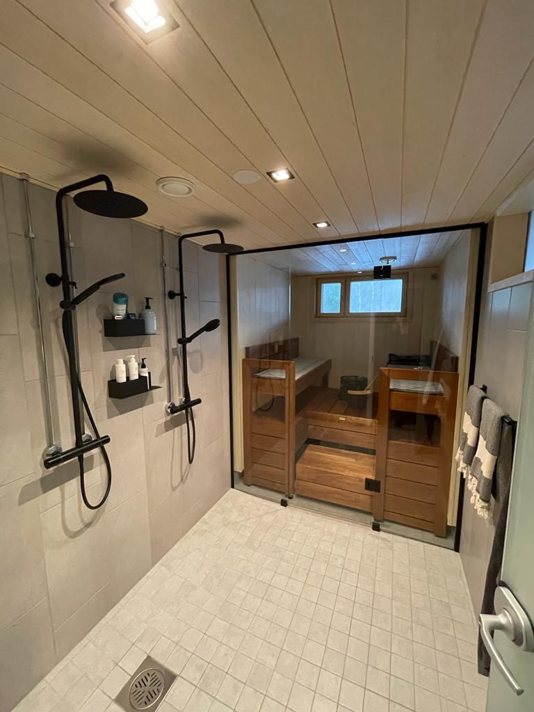 Moderni suihkuhuone, jossa valkoiset laatat, mustat kalusteet ja lasiovien läpi näkyvä puinen sauna