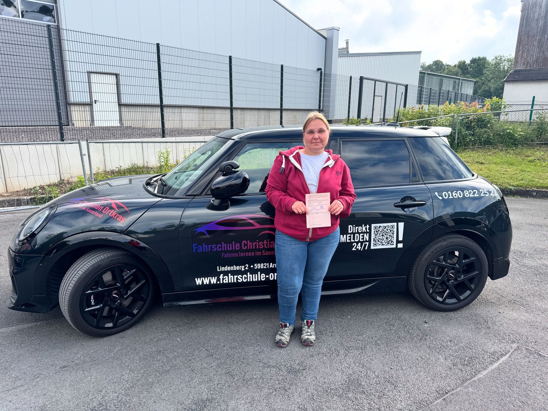 Eine Frau steht mit einer Urkunde vor einem schwarzen Auto.