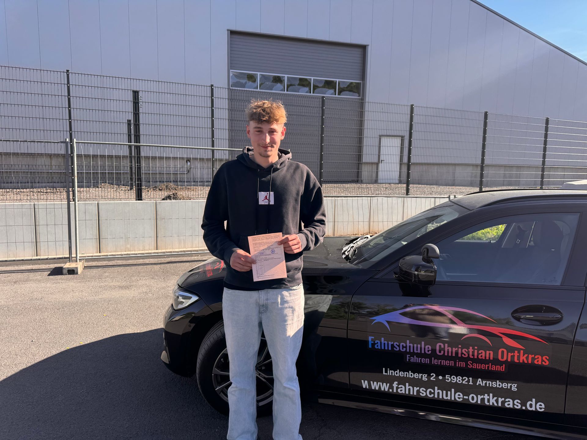 Ein junger Mann steht mit einem Zertifikat vor einem schwarzen Auto.