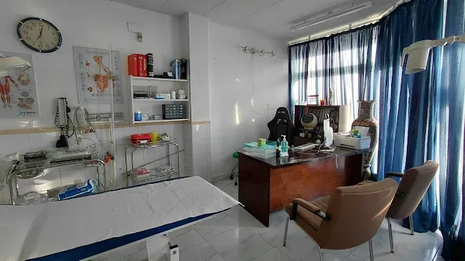 Habitación de hospital luminosa con cama, escritorio, sillas, estantes y cortinas azules junto a la ventana.