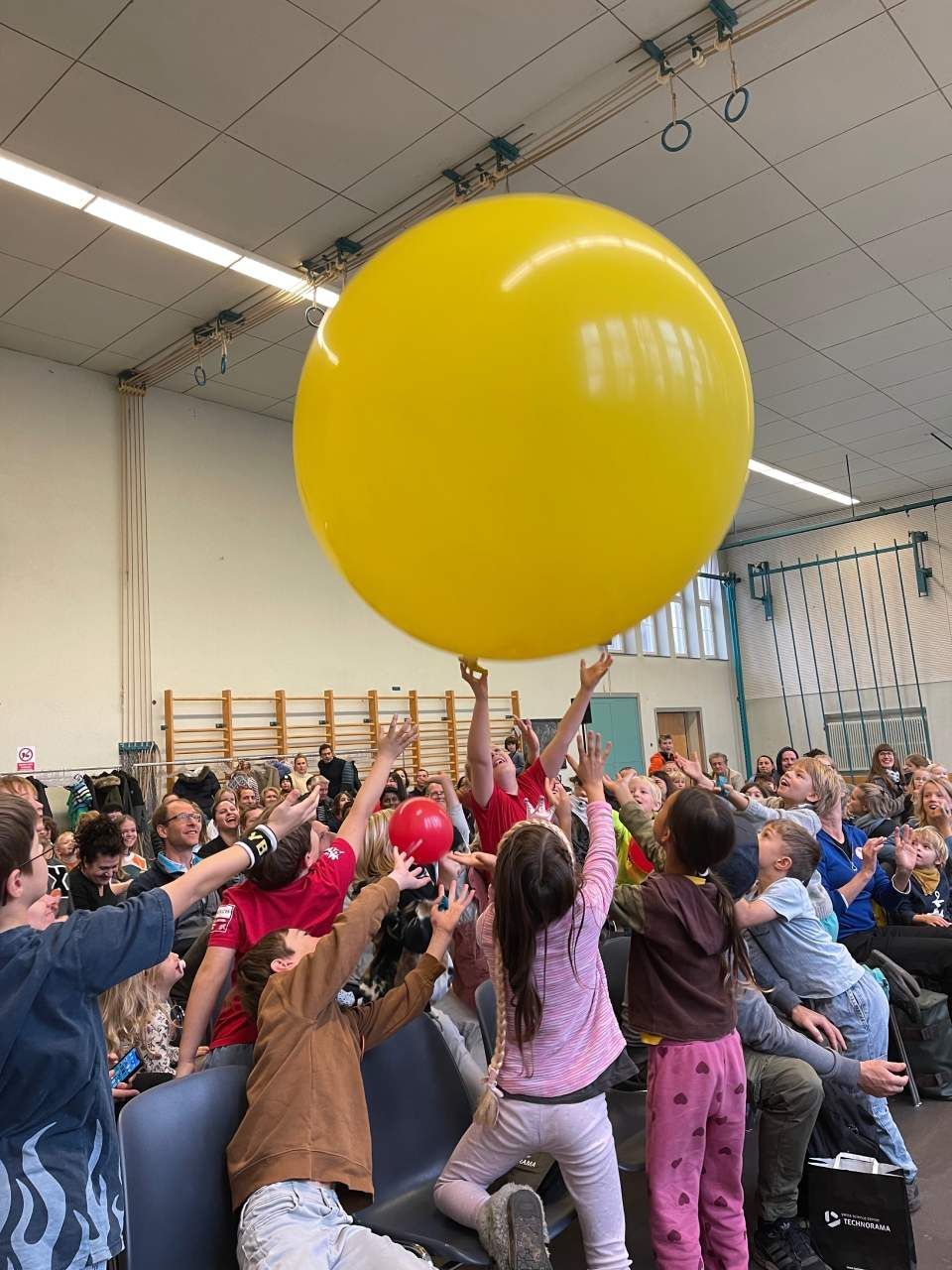 Kinder greifen in einem Raum nach einem grossen gelben Ballon. Andere Kinder schauen zu.

