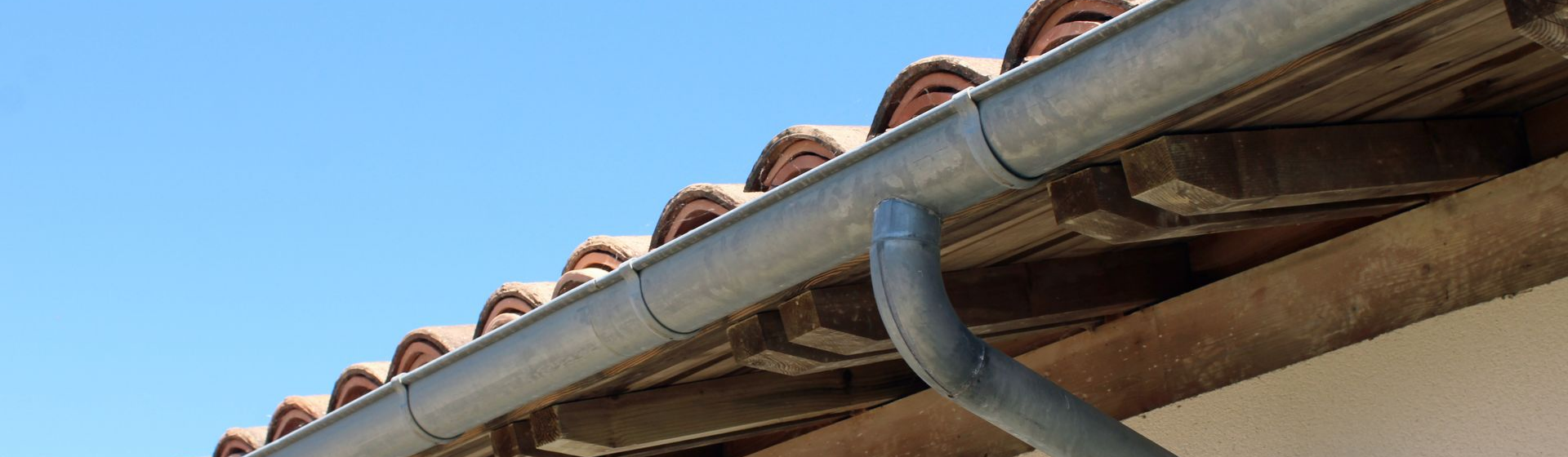 Une gouttière métallique avec une descente pluviale sur un toit de tuiles en terre cuite, se détachant sur un ciel bleu.