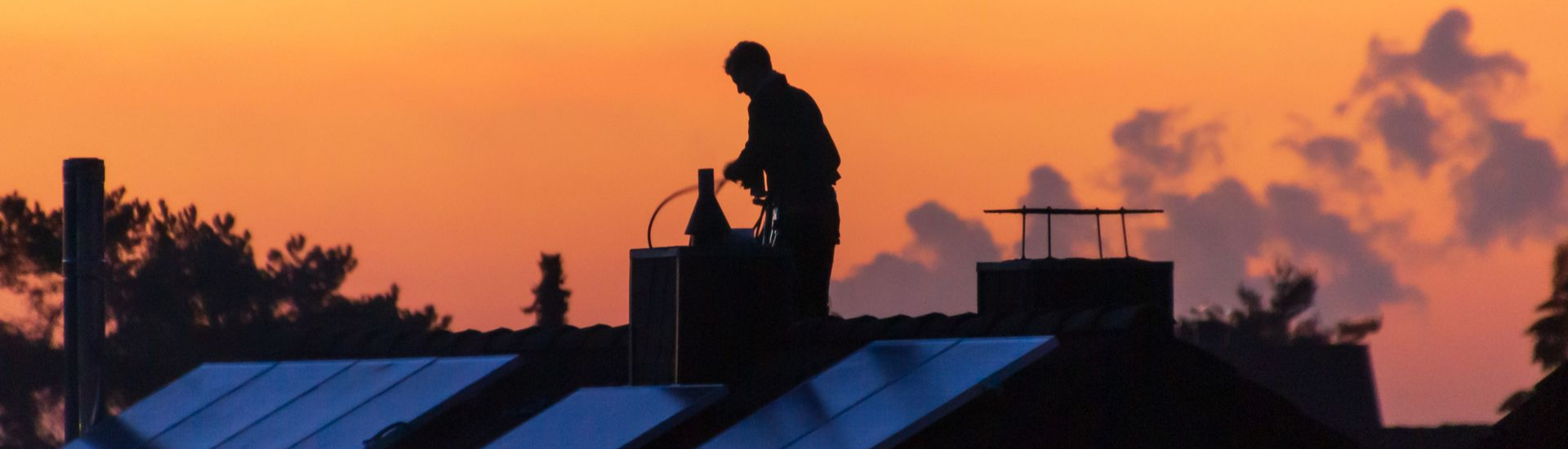 Silhouette d'une personne sur un toit avec une cheminée, se détachant sur un coucher de soleil orangé et bleu.