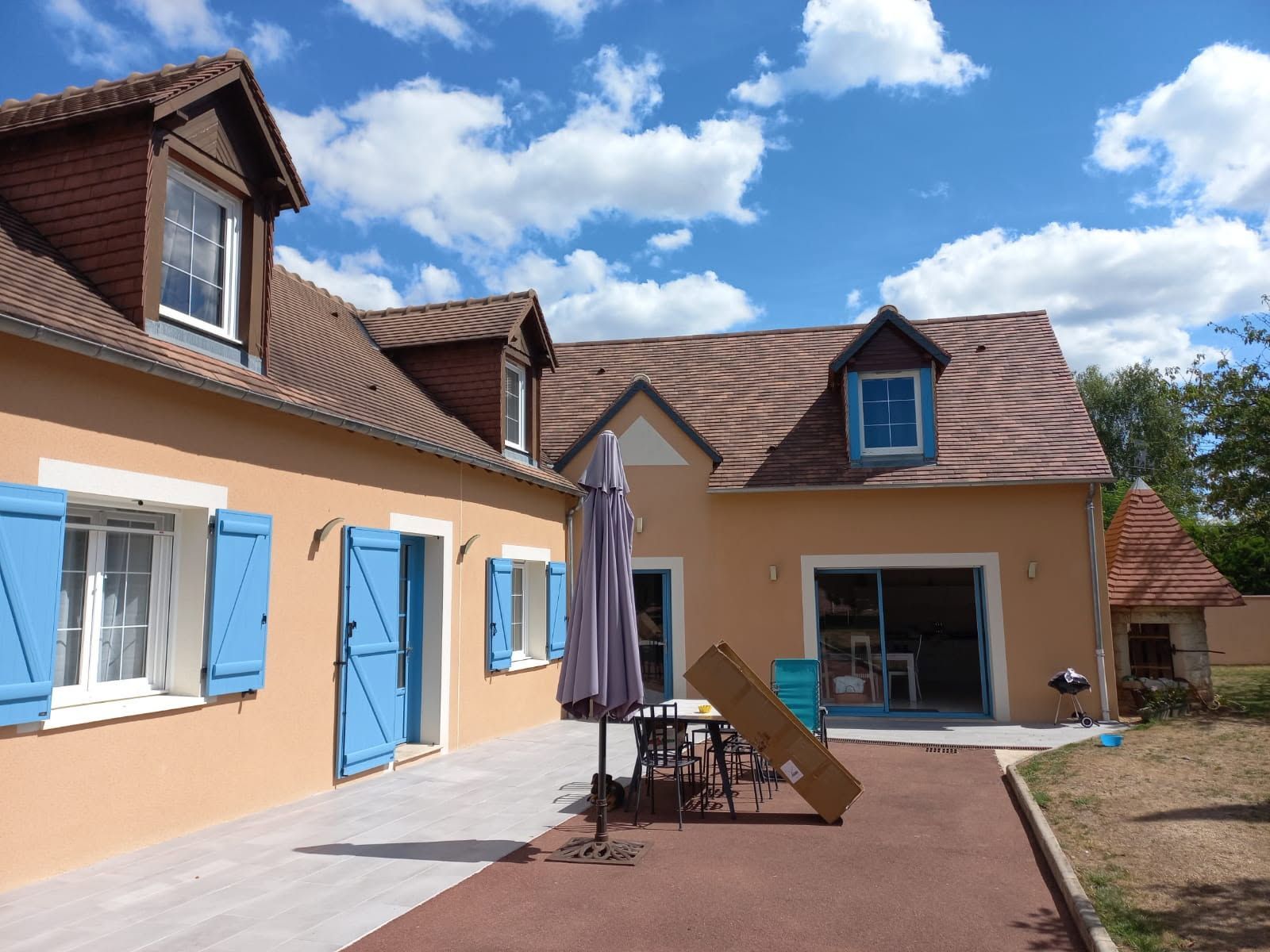 Maison couleur pêche avec volets bleus, toit marron, lucarnes et patio par une journée ensoleillée.
