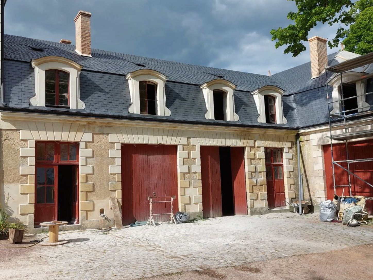 Bâtiment en pierre au toit gris foncé, aux portes et fenêtres rouges, sous un ciel nuageux.