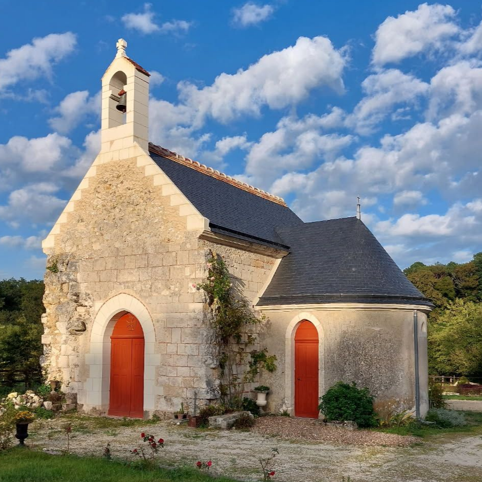 Petite chapelle en pierre aux portes rouges et au clocher, sous un ciel bleu nuageux.