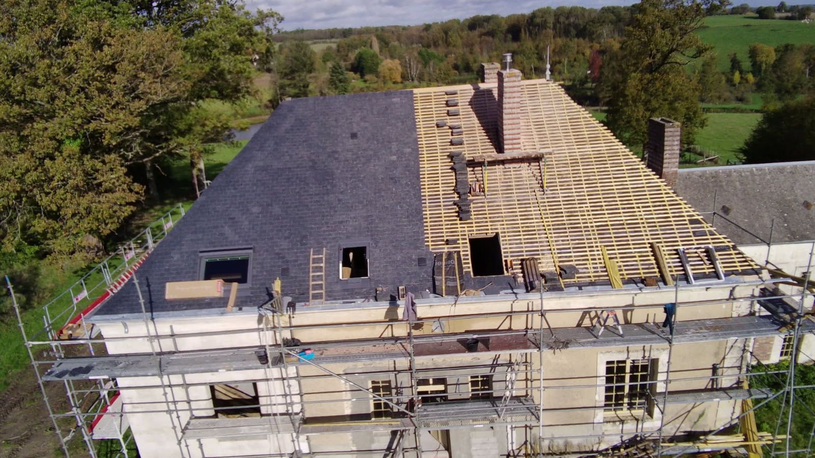 Toiture de la maison partiellement recouverte de bardeaux gris foncé, l'autre moitié laissant apparaître les chevrons en bois, échafaudages sur la façade.