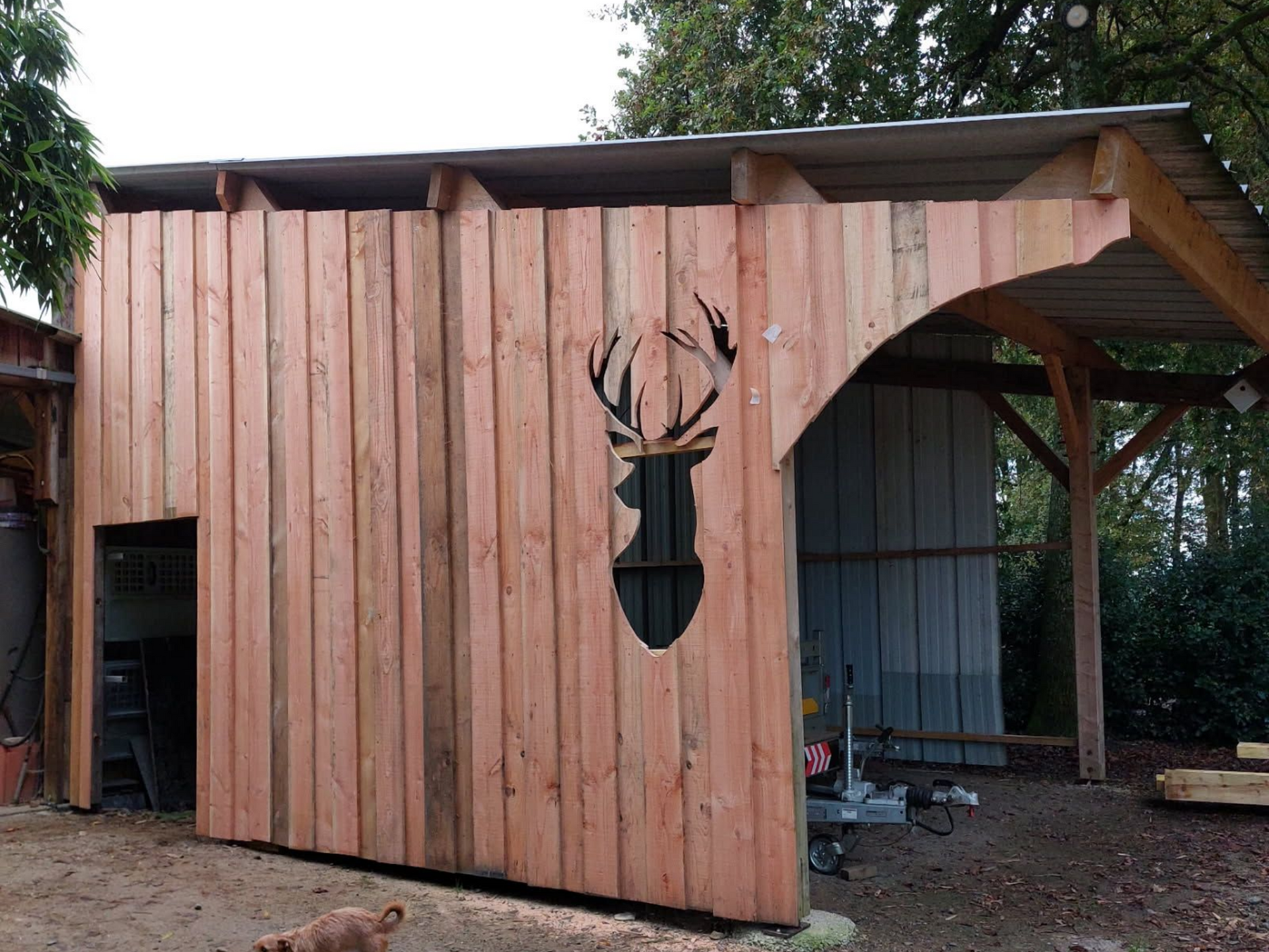 Abri en bois avec découpe en forme de tête de cerf, toit en métal, côté ouvert avec machines, aménagement extérieur.