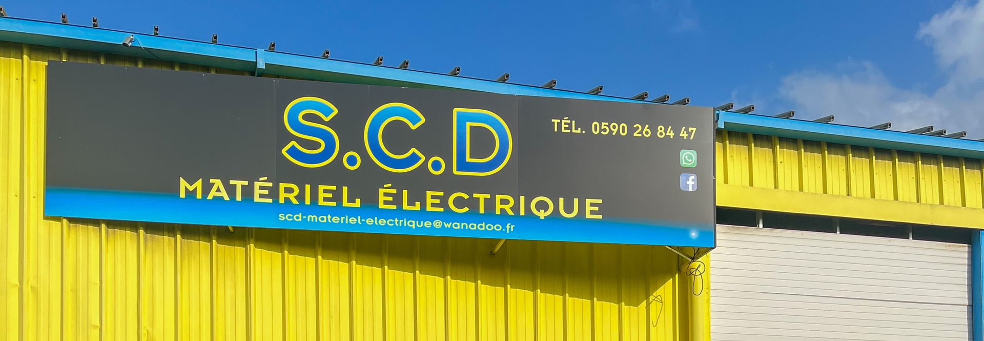 Façade du magasin SCD Matériel Électrique avec extérieur jaune et gris sous un ciel bleu clair