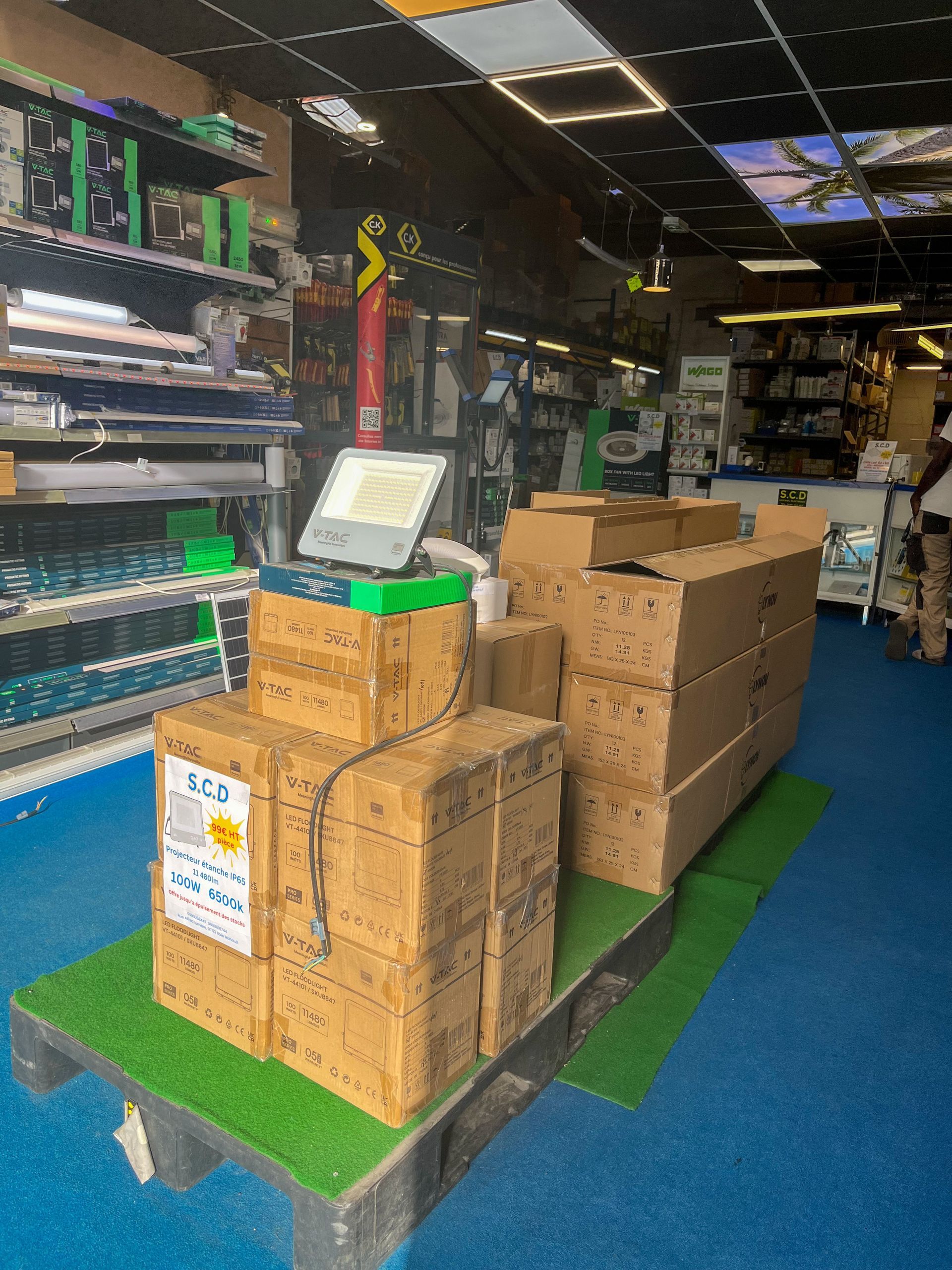 Des cartons empilés sur des palettes vertes dans une allée de magasin au sol bleu, avec des étagères en arrière-plan.