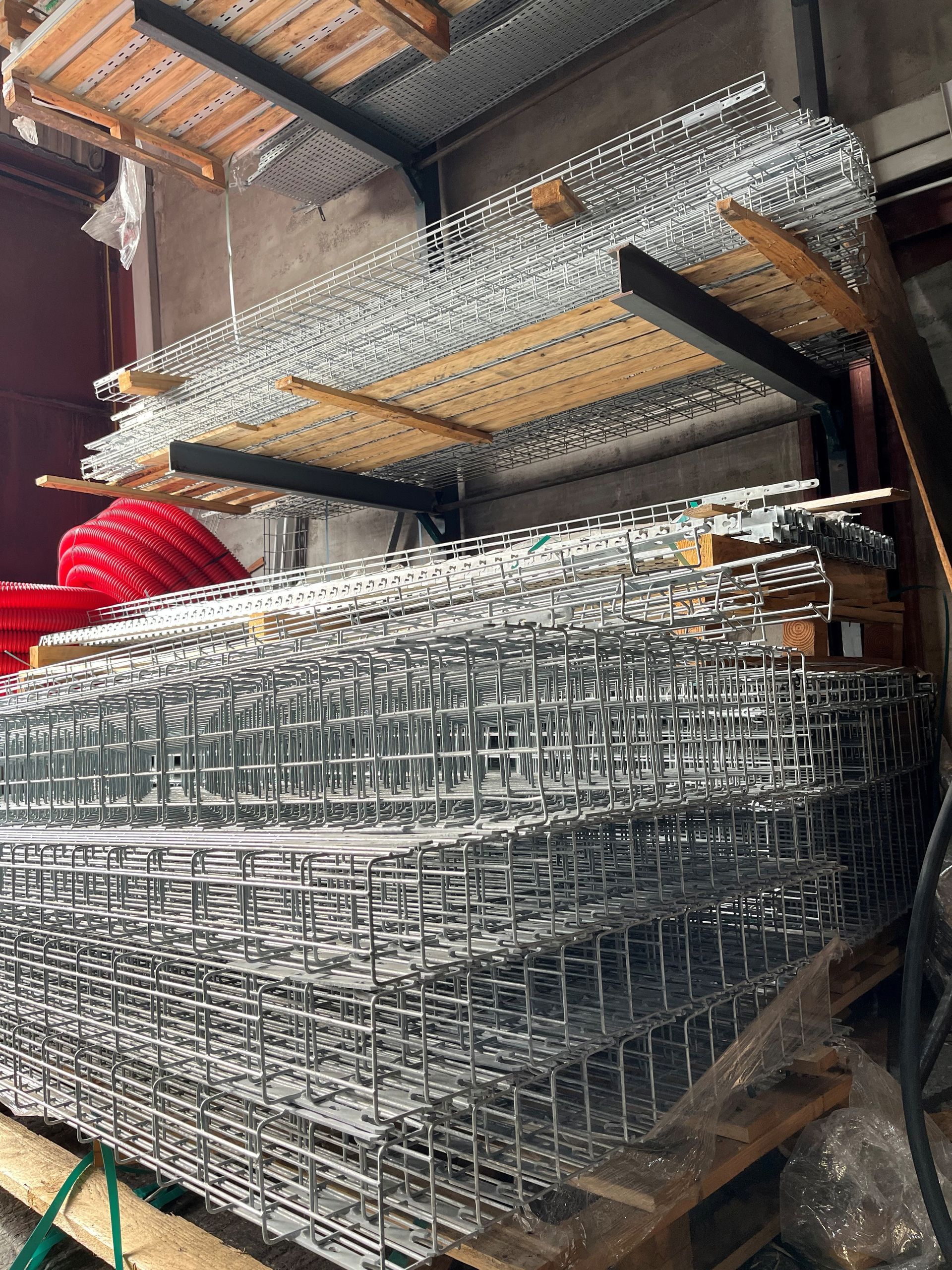 Des cages en grillage empilées dans un entrepôt, avec des étagères et un objet rouge en arrière-plan.