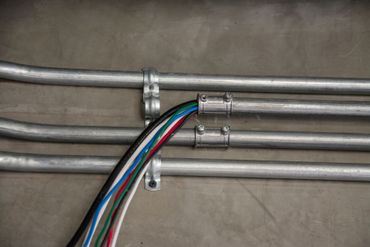 Des fils électriques colorés, regroupés, sont fixés à un conduit métallique contre un mur en béton.