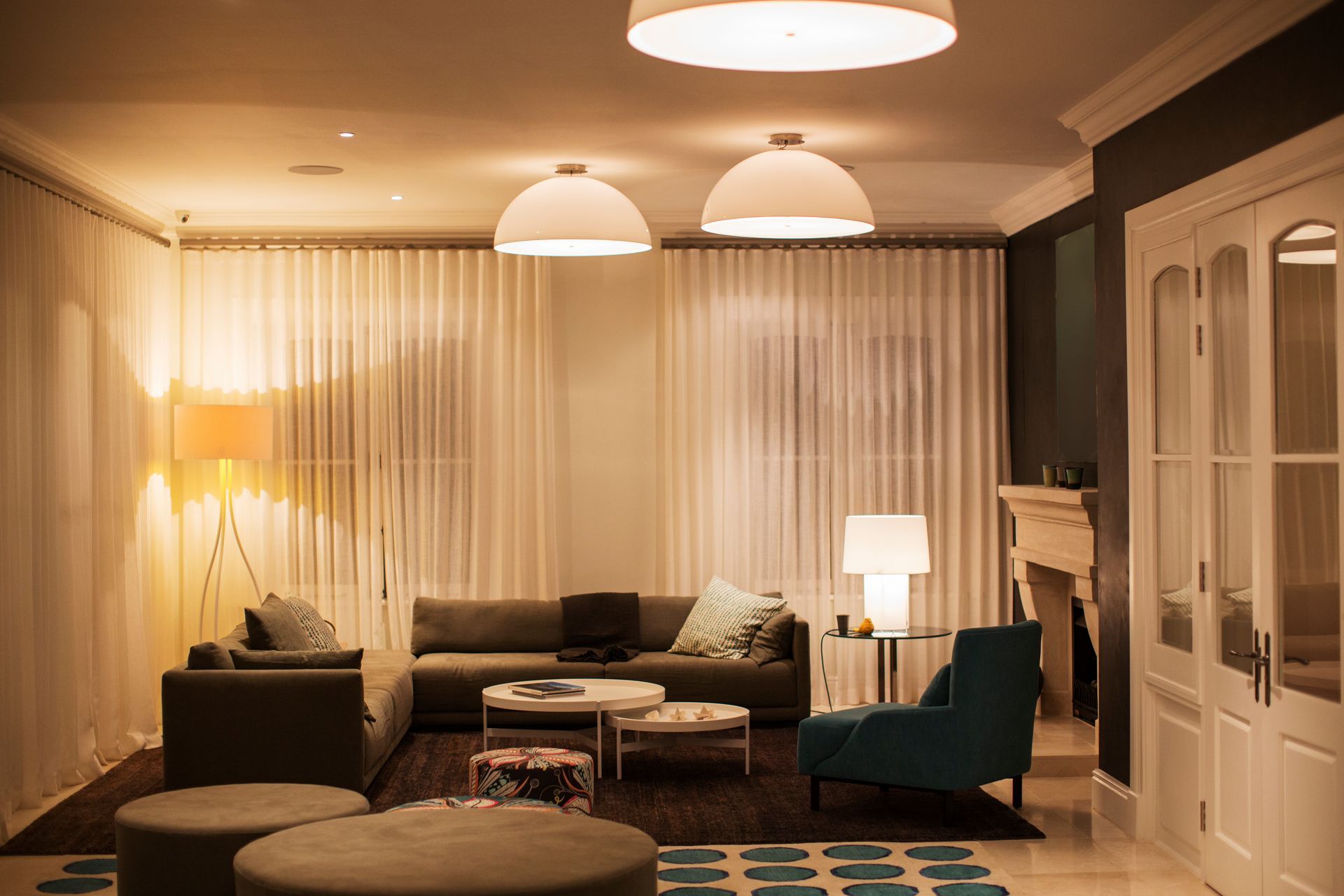 Salon confortable avec canapé, fauteuil turquoise, lampadaire, table basse ronde et rideaux légers sous une lumière chaude.