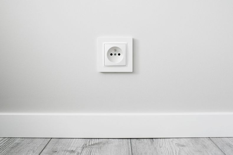 Mur blanc avec une prise électrique centrée au-dessus d'un parquet clair et de plinthes.