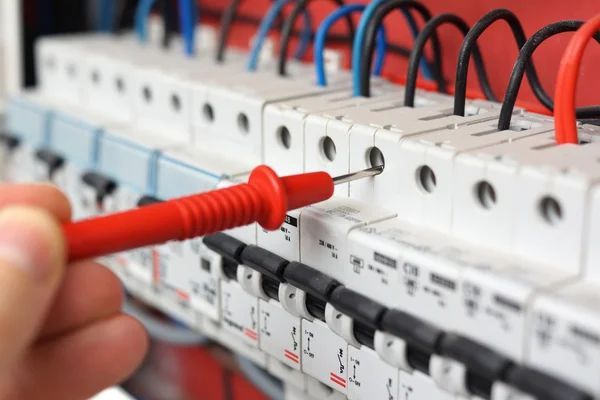 Une sonde rouge teste une rangée de disjoncteurs électriques avec des fils bleus et rouges.