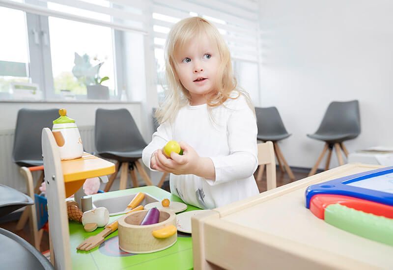 Ein Kind hält eine gelbe Frucht an einem Spieltisch in einem hellen Klassenzimmer, in dessen Nähe sich Spielzeug und Stühle befinden.