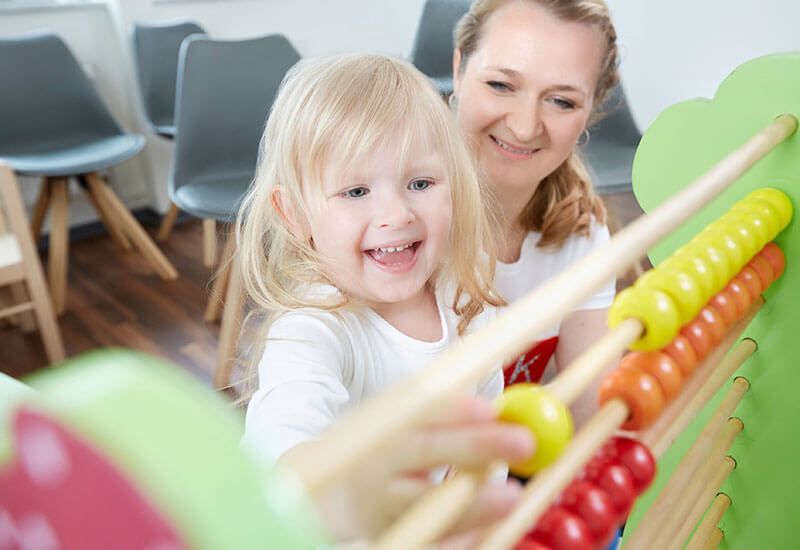 Frau und lächelndes Kind spielen in einem hellen Raum mit einem bunten Abakus.