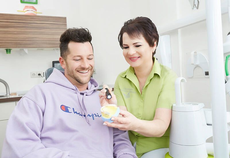 Ein lächelnder Mann und eine lächelnde Frau zeigen in einer Zahnarztpraxis einem Patienten auf dem Stuhl eine Babyflasche.