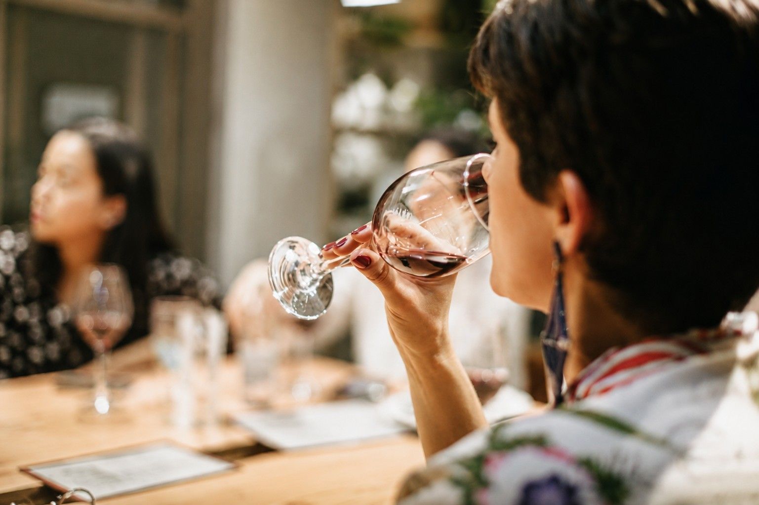 Une personne sirote un verre de vin à une table de restaurant tandis que d'autres discutent en arrière-plan.