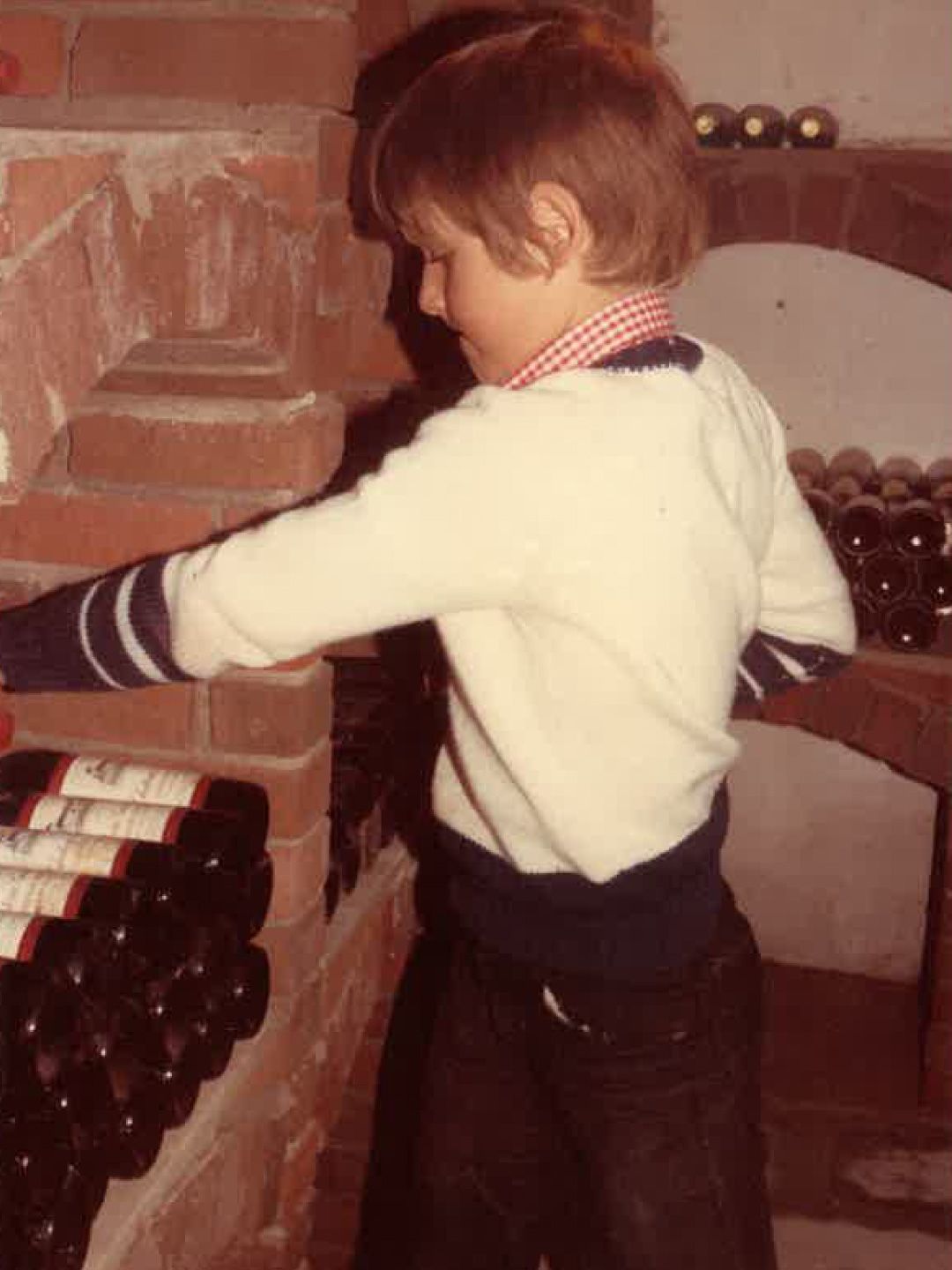 Un enfant tend la main vers des bouteilles posées sur une étagère à côté d'une cheminée en briques.