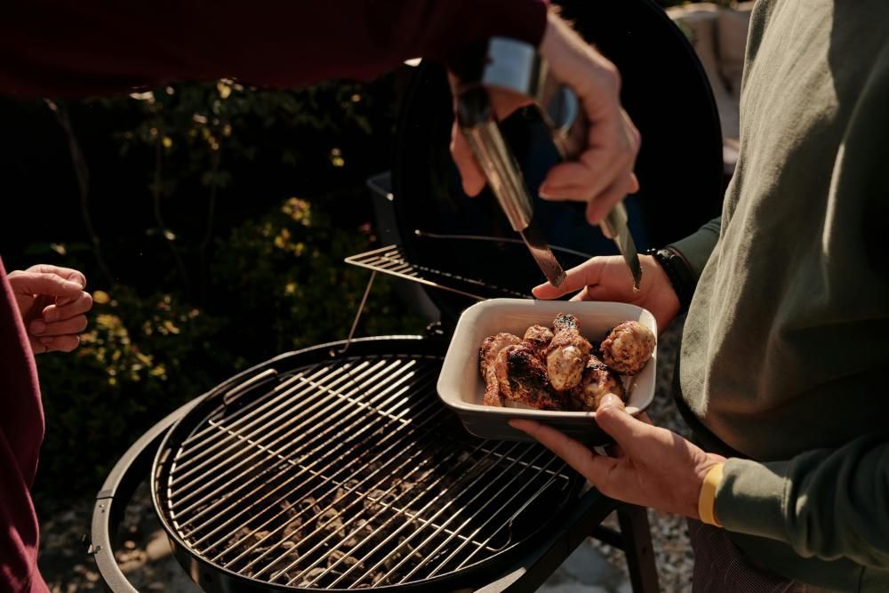 Des personnes servent du poulet grillé lors d'un barbecue, avec un gril et une barquette en aluminium à l'extérieur.