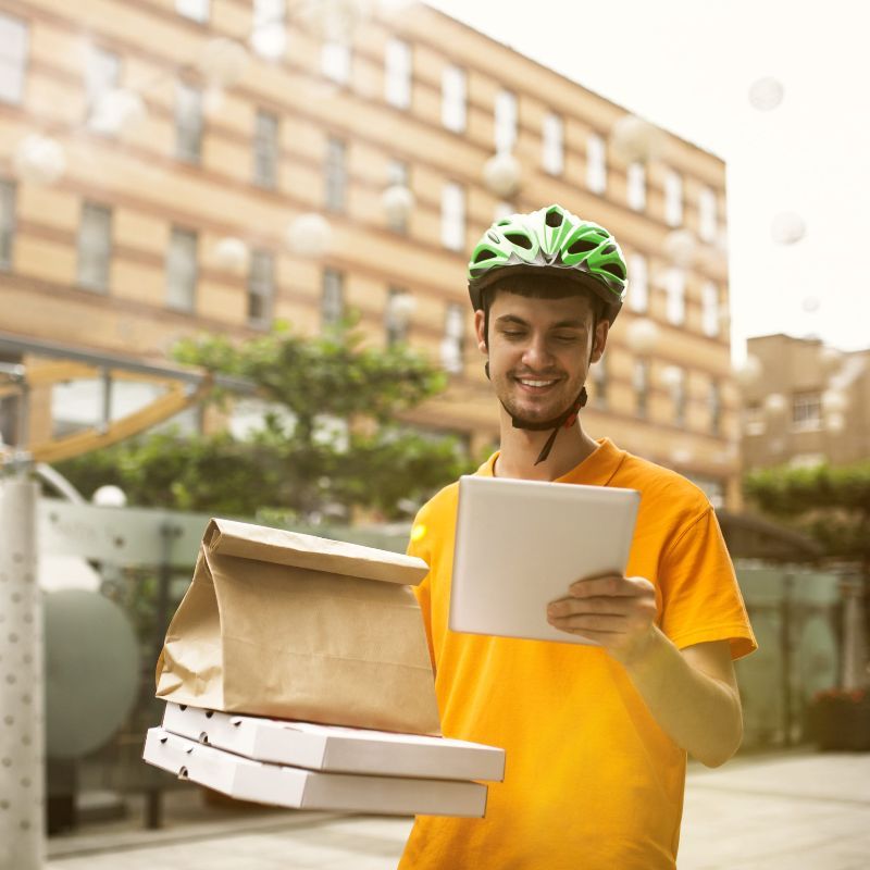 Un repartidor de comida, vestido con una camisa amarilla y un casco verde, lleva dos cajas de pizza, una bolsa de papel