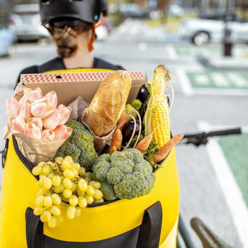 Un repartidor de comida en bicicleta lleva una mochila amarilla brillante repleta de productos frescos, pan y flores.