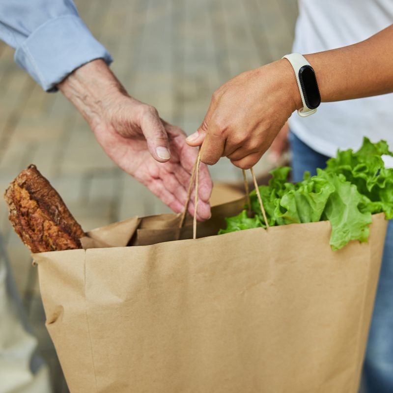 Dos personas intercambian una bolsa de papel de la compra llena de lechuga fresca y una barra de pan.