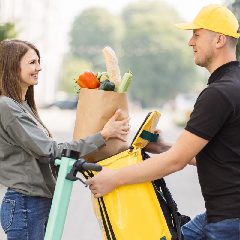 Un repartidor con gorra amarilla entrega una bolsa de papel con productos frescos a un cliente que está de pie