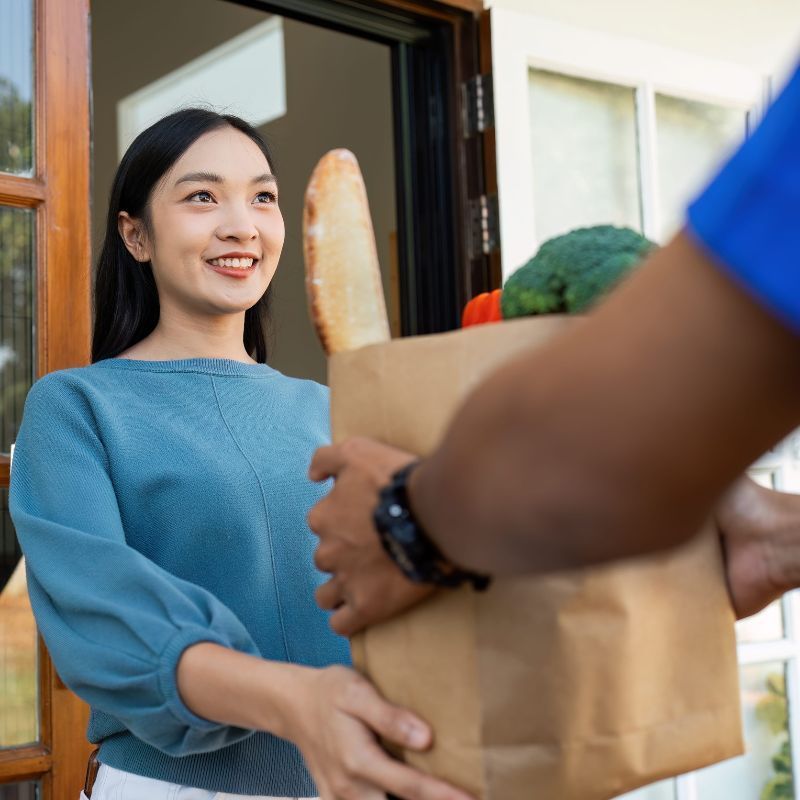 Una persona le entrega una bolsa de papel marrón llena de productos a otra persona en la puerta de una casa.