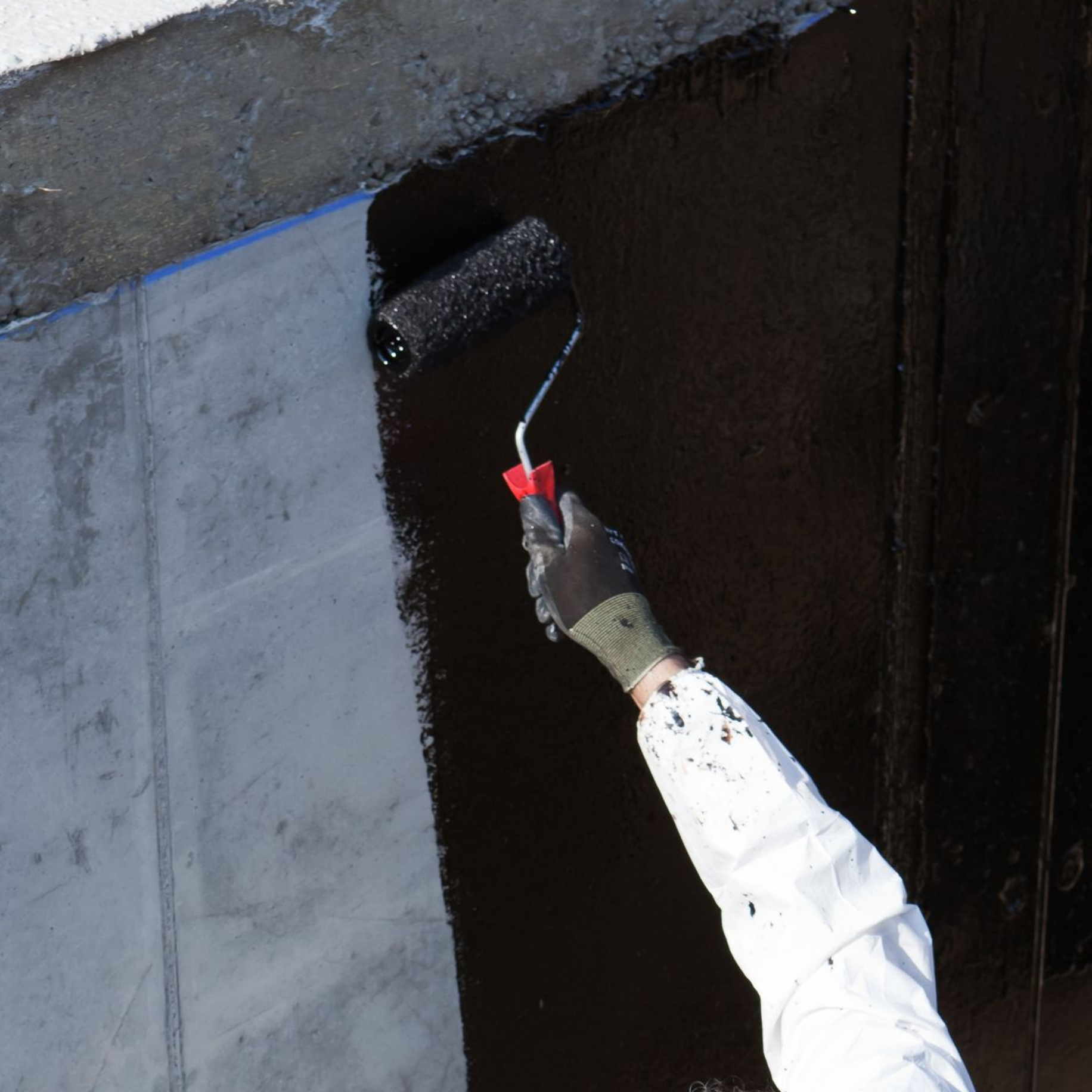 Une personne peint un mur en béton au rouleau, appliquant une couche foncée pour préserver l'étanchéité d'un mur enterré