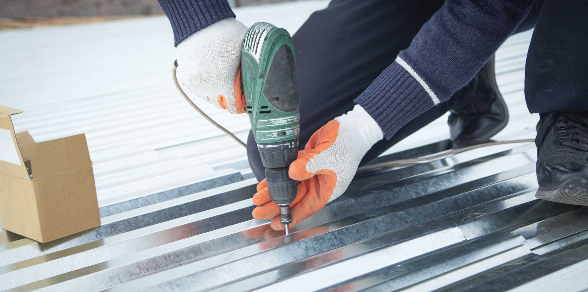 Des mains gantées utilisent une perceuse verte pour fixer des panneaux de toiture métalliques sur un toit